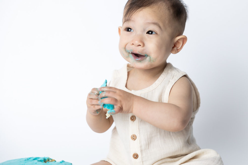 First Birthday Cake Smash photography - baby looking very happy eating cake by Paper Bunny Studios Edmonton