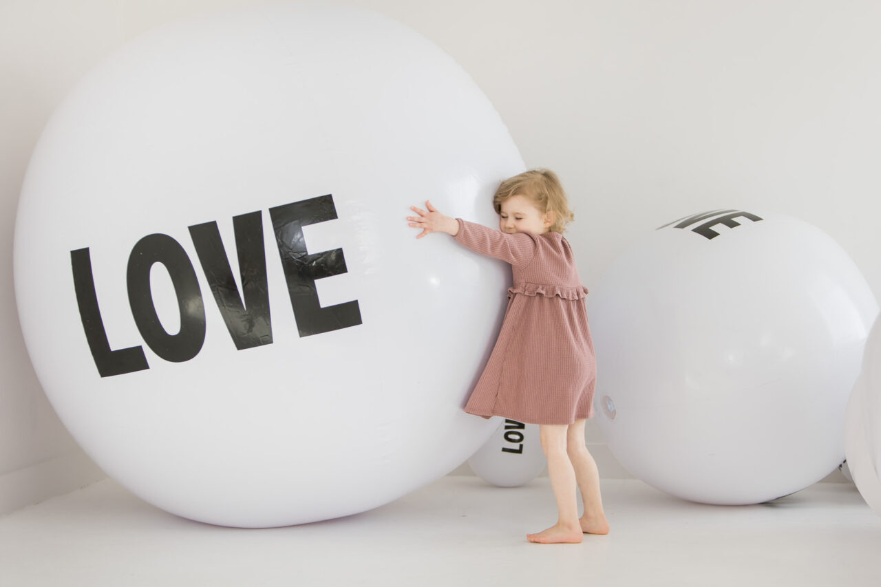 Young girl hugging a beach ball that says LOVE on it during LOVE mini session with Paper Bunny Studios using giant beach balls saying LOVE on them as a backdrop. Image copyright Paper Bunny Studios